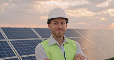 Portrait of happy male engineer in protective helmet crossing arms while looking to camera. Handsome man in uniform smiling while standing at solar power farm. Concept of green energy