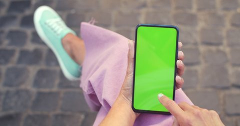 Crop view of female person holding and scrolling smartphone with mockup screen while sitting at street. Concept of chroma key and greenscreen. Paving stones background