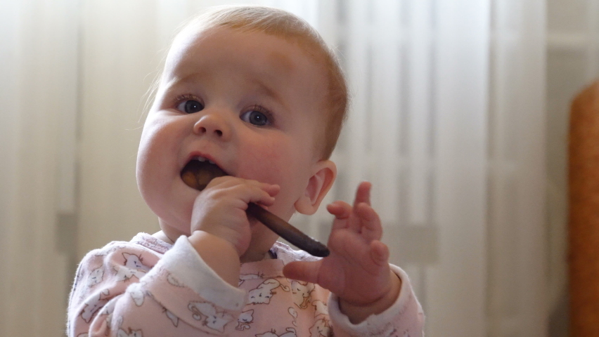 The kid chews a wooden spoon. The child plays on the floor in the room. They cut their teeth. Baby. Daylight. Close-up.