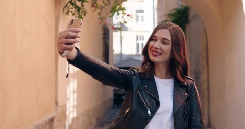 Beautiful joyful young Caucasian woman in black leather jacket posing to smartphone camera and taking selfie photo. Pretty stylish female tourist making photos with mobile phone at cozy street.