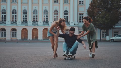 Brunette guy sitting on longboard and his female friends pushing him forward and running across the street. Teenage friends having fun together. Adventurous teen spirit ans active sport life.