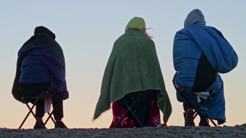 A group of young friends meet the sunrise on the mountain. Sleepy tourists in sleeping bags, sitting on chairs on a cliff. A magnificent view of the city at the foot of the mountain