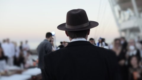 A man with a black hat on his head walks among other people at the beach club. The camera captures the man from the back.