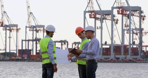 Seaport workers work with a paper drawing against the background of cargo cranes. Engineers discuss with a worker the cargo logistics improvement at the port.