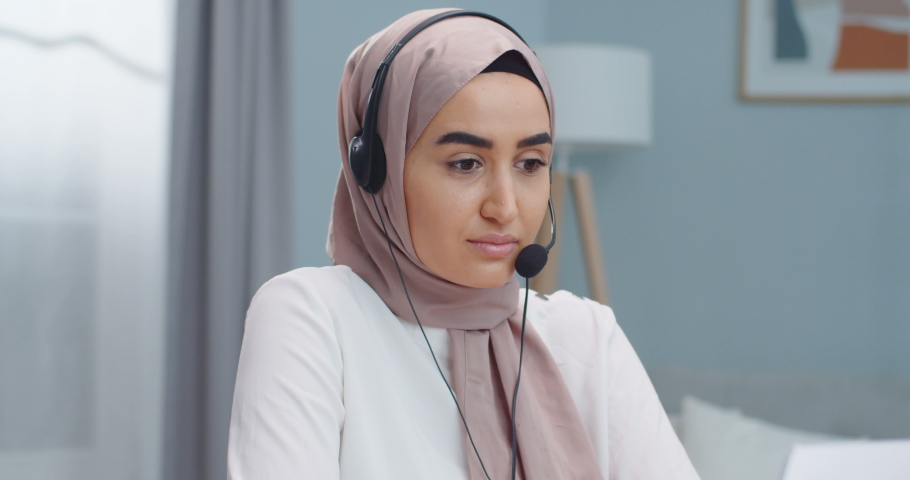 Emotional young muslim woman sales representative. Female in hijab and headsets speaking to client, making video conference call, looking at laptop, talking by webcam, sell online, work from home. Royalty-Free Stock Footage #1059478988