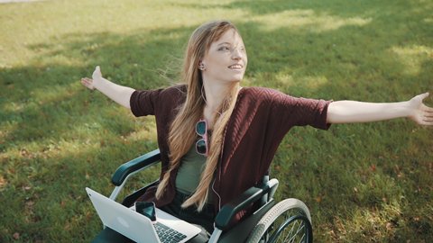 Young disabled caucasian woman in the wheelchair with laptop enjoying the autumn day in the park with music from smartphone. High quality 4k footage