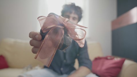 Young adult male holding surprise in hand in the act of giving a gift in video call to distant partner. Distant couple sharing presents in video conference, looking in camera  bright home interior .