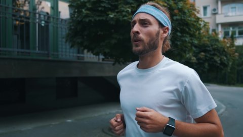 Sporty young bearded man runs on streets and checks time on smartwatch. Hipster guy wearing headband enjoying morning run in the city.