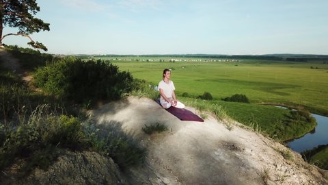 Aerial shot view of yogini young woman sitting in lotus pose and putting up hands in namaste mudra. Female practicing yoga on top of rock background of flowing river and green meadow on summer day.