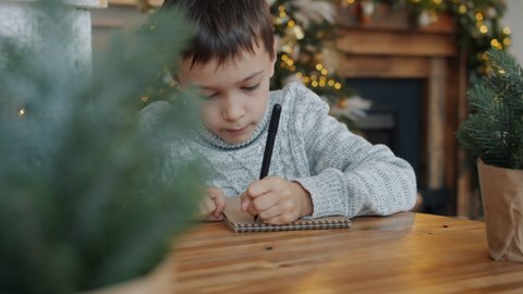 Portrait of thoughtful little boy writing letter to Santa Claus before Christmas in decorated room, child is sitting at table alone. New Year and holidays concept.