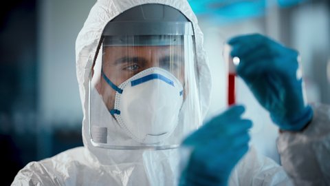 Medical Research Scientist Wearing Coverall, Surgical Gloves, Face Mask and Shield Holds Test Tube with Blood Sample and Label Reading Covid-19. Microbiology Laboratory Drug and Vaccine Development