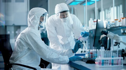 Medical Laboratory: Team of Microbiology Scientists Wearing Sterile Coveralls, Face Shields and Masks Talk, Use Computer to Analyse Test Tube Blood Samples and Develop Vaccine, Drugs and Antibiotics