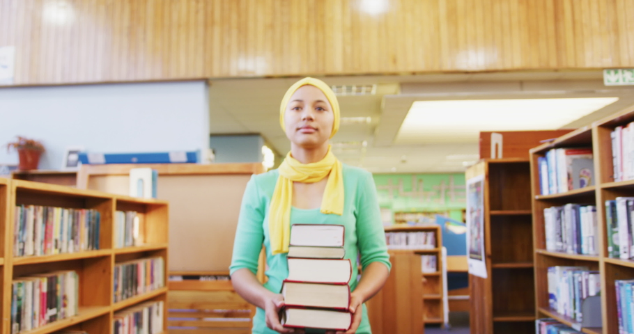 An Asian female student wearing a yellow hijab studying in a library, walking between bookshelves and holding a pile of books, wearing casual clothes