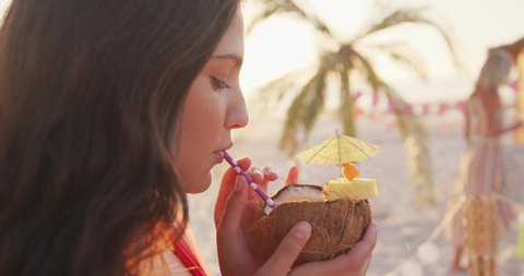 Side view of a Caucasian woman on holiday enjoying time together on a tropical beach at sundown, drinking and smiling, in slow motion