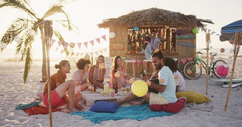 Front view of mixed race friends on holiday enjoying time together on a tropical beach at sundown, sitting at the table and laughing, in slow motion