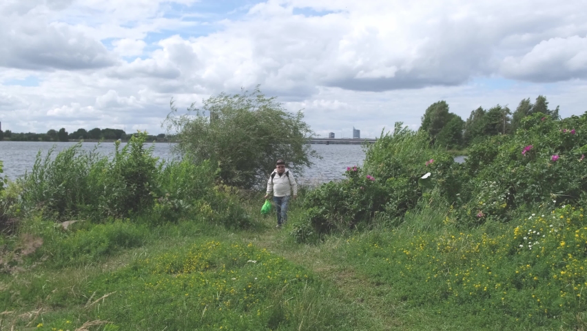 Mature man in hat and white sweatshirt with backpack walks along narrow rural path on riverbank on windy summer day