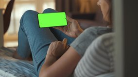 Woman at home lying on a sofa and using smartphone with green mock-up screen in horizontal mode. Girl browsing Internet, watching content, videos, blogs.