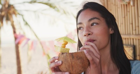 Close up of attractive mixed race woman on holiday sitting at a bar on a tropical beach in the sun smiling and drinking a cocktail from a coconit shell, in slow motion. Relaxing tropical beach holiday