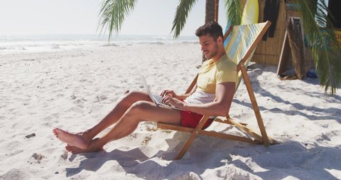 Caucasian man enjoying time at the beach, sitting on a sunbed and using his laptop on a sunny day in slow motion. Relaxing Summer Beach Vacation.