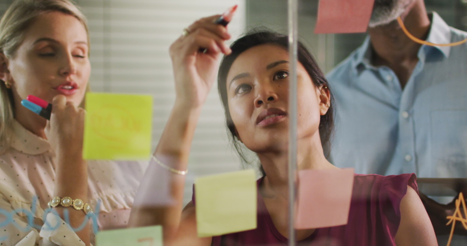 Caucasian and Asian businesswomen and mixed race businessman working late in the evening in a modern office together, drawing on glass moodboard with memo notes, brainstorming in slow motion. | Shutterstock HD Video #1059504350