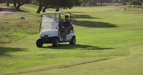 Two Caucasian male golfers playing a game, driving in a golf buggy on a golf course on a sunny day wearing caps and golf clothes, their clubs in the back , in slow motion 