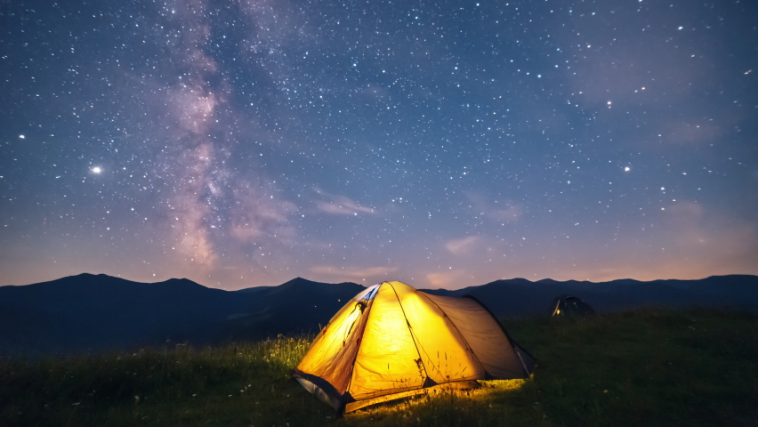Camp tent with a burning light inside under the starry sky and rising Milky way Galaxy over the high mountains, night landscape