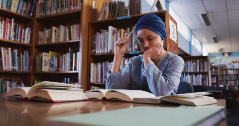 An Asian female student wearing a blue hijab studying in a library, sitting at a desk with open books and taking notes, wearing casual clothes in slow motion