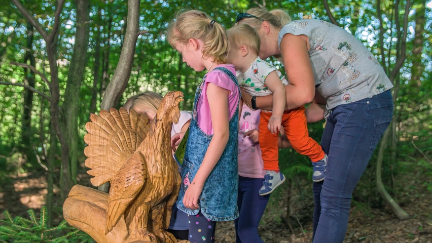 Children and mom exploring wooden statue of a peacock placed inside green park surrounded by lush vegetation, slow-mo shot