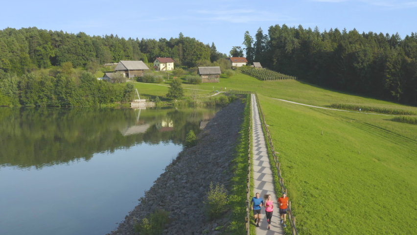 Aerial footage of two man and one woman running on the track fenced with wooden fence by the lake, Fitness and recreation concept
