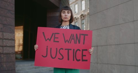 Millennial woman holding carton banner with we want justice writing on it. Hipster female with nose ring supporting human rights movement while standing at empty street. Zoom in.