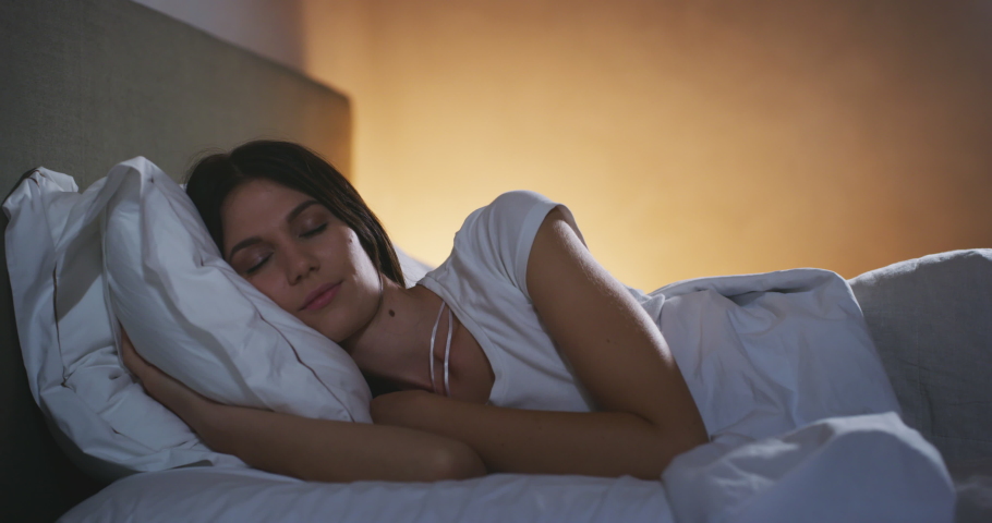An young brunette woman is sleeping peacefully under warm duvet blanket in a cosy bed with switched on night lamp in a bedroom. Concept of comfort, relax,sleeping,health, bedding, softness,dreams Royalty-Free Stock Footage #1059525389