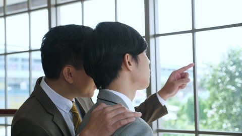 Medium shot of middle ages asian businessman in formal suit walks in, greets the junior by touching the shoulder, giving compliment to cheer up junior man who is looking out throught the windows