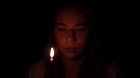 Woman is blowing off the candle at black background and disappears into darkness. Portrait of young caucasian lady standing in the studio with burning candle. Theme of magic and mystical rituals.