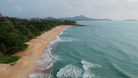 Aerial Drone Shot. Flying along the ocean coast. View of the sea waves, sandy beach, green hill, blue water and palm trees. The island Koh Samui, Thailand. Near Samui Resort Hotel. Full HD video.