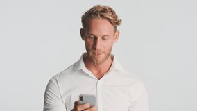 Attractive serious blond bearded man in shirt intently using smartphone waving no gesture on camera over white background