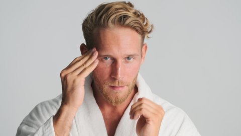 Attractive blond bearded man in bathrobe applying cream on face and smiling on camera over white background