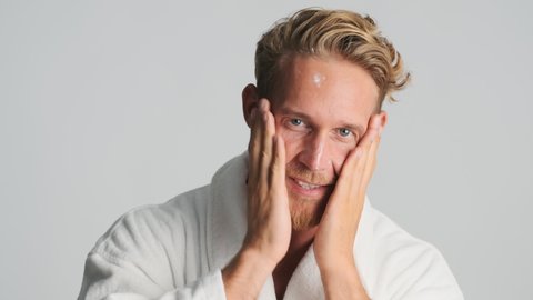 Handsome blond bearded man in bathrobe happily applying cream on face over white background