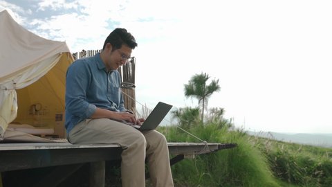 Young attractive happy asian guy using laptop computer work remotely anywhere using internet connected to office. Freelancer male work on vacation with scenic view of mountain, forest and tent resort.
