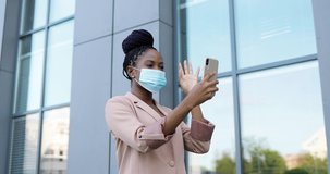 Pretty happy African American young woman in medical mask having videochat on smartphone outdoor at business building. Cheerful beautiful female talking and videochatting via webcam on mobile phone.