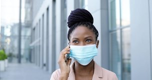 African American young beautiful cheerful woman in medical mask talking on mobile phone and laughing outside at business center. Happy businesswoman in respiratory protection speaking on cellphone.