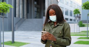 African American young stylish pretty woman in medical mask standing at urban street and using smartphone. Beautiful female outdoors in city texting message on mobile phone. Coronavirus pandemic.