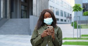 African American young stylish woman in medical mask using smartphone, tapping and scrolling on screen. Beautiful female outdoors in city texting message on mobile phone. Coronavirus. Urban street.
