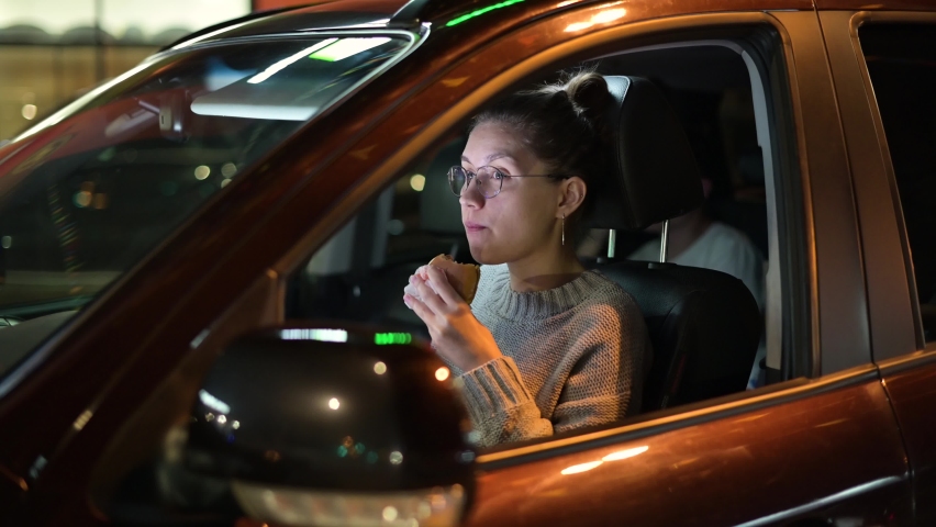 The camera zooms in on a young woman eating a burger while sitting in her car in a parking lot in the evening. 