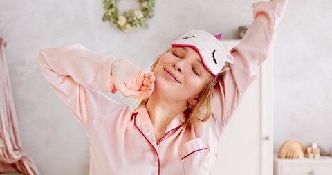 Young yawns woman in pink pajamas and sleep mask, waking up and stretching after sleep, sleepy and happy. Good and healthy sleep concept.