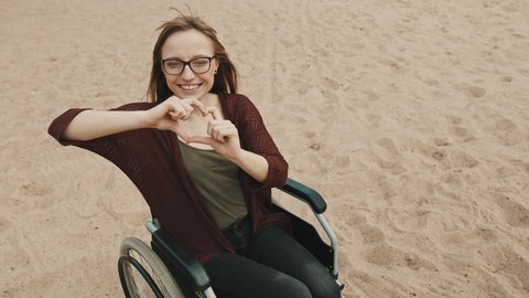 Suport for disabled people. Young woman in the wheelchair on the sandy river coast showing heart with hands. High quality 4k footage