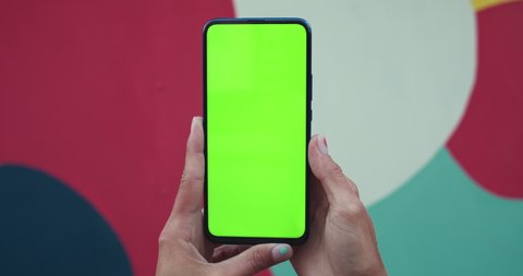 Close up view of female hands with trendy fingernails design holding smartphone with mockup screen at colourful spots background. Concept of green screen and chroma key