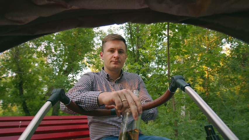 The child sees the father drinking alcohol. A newborn child looks from a pram at his father sitting on a Park bench and drinking beer from a transparent glass bottle, a view from a pram from the child