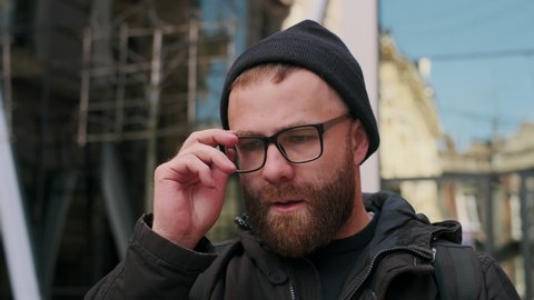 Portrait of bearded guy taking off his glasses and thinking while walking at city street. Close up view of sad male person having bad news and looking thoughtful. Concept of emotions
