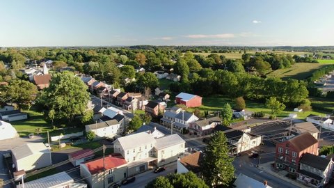 High and wide aerial overhead shot of small American town, village, establishing shot shows homes and housing along street, green trees and farmland in distance during August, North America, USA