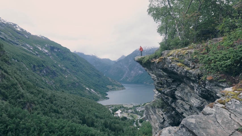 Person standing on the edge of a mountain, throwing arms up into the air, surrounded by nature, mountains and watching over a beautiful lake. Norway Geiranger